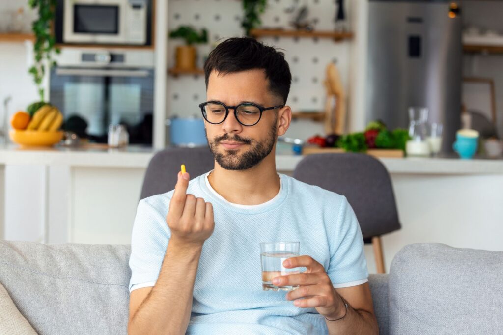 A man holds a pill and a glass of water, sitting on a sofa