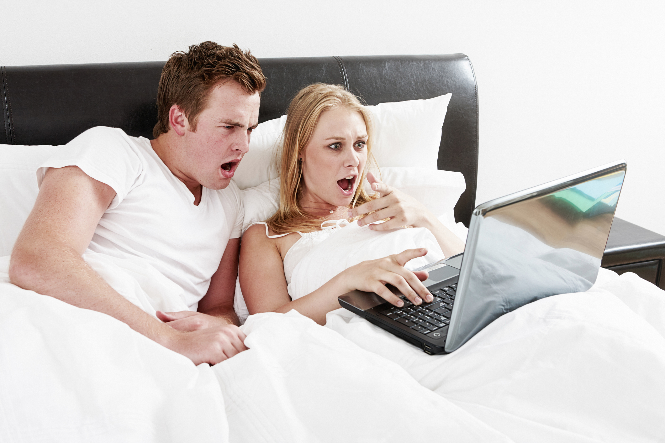 Scandal! Shocked young couple react in shock and horror to their laptop computer as they lie in bed togther