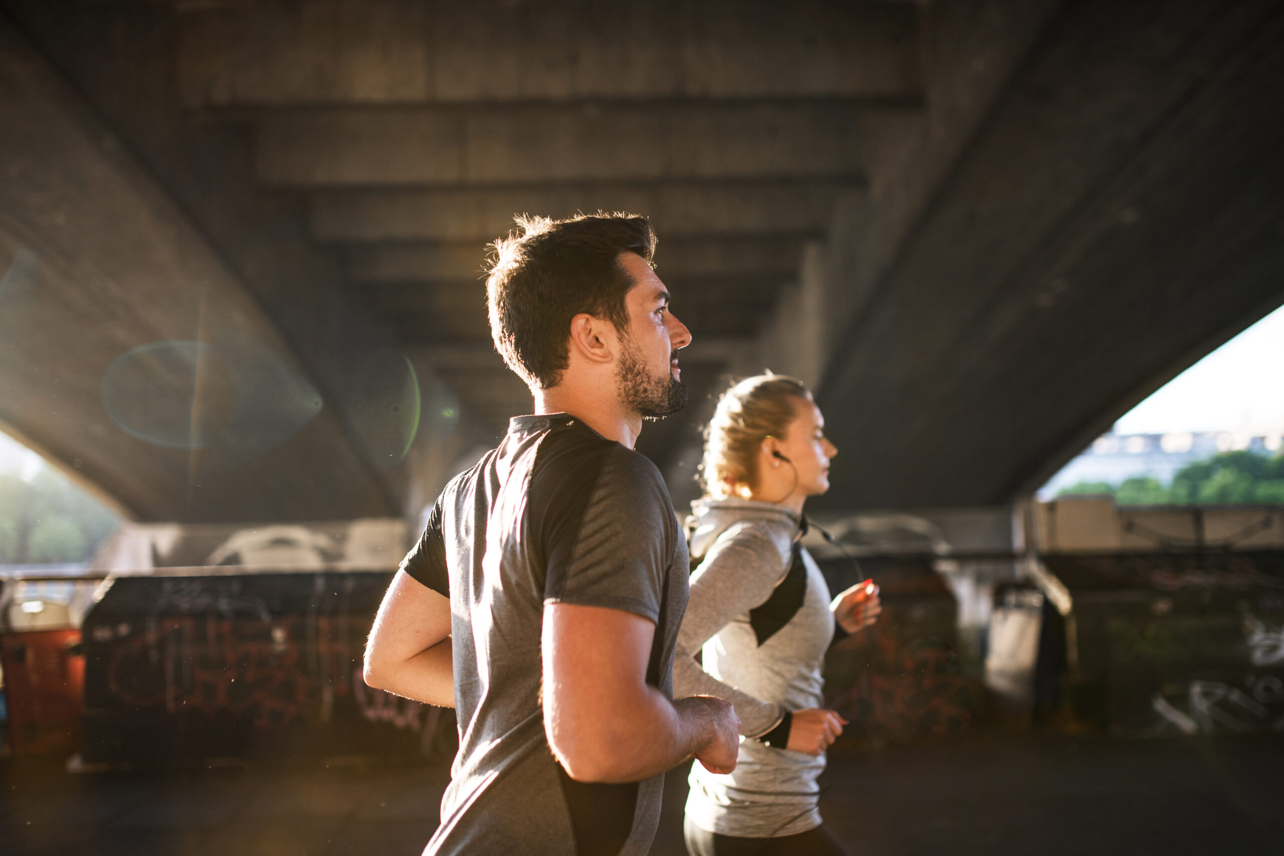 Young sporty man and woman with earphones running under the bridge outside in a city. Jogger's nipple