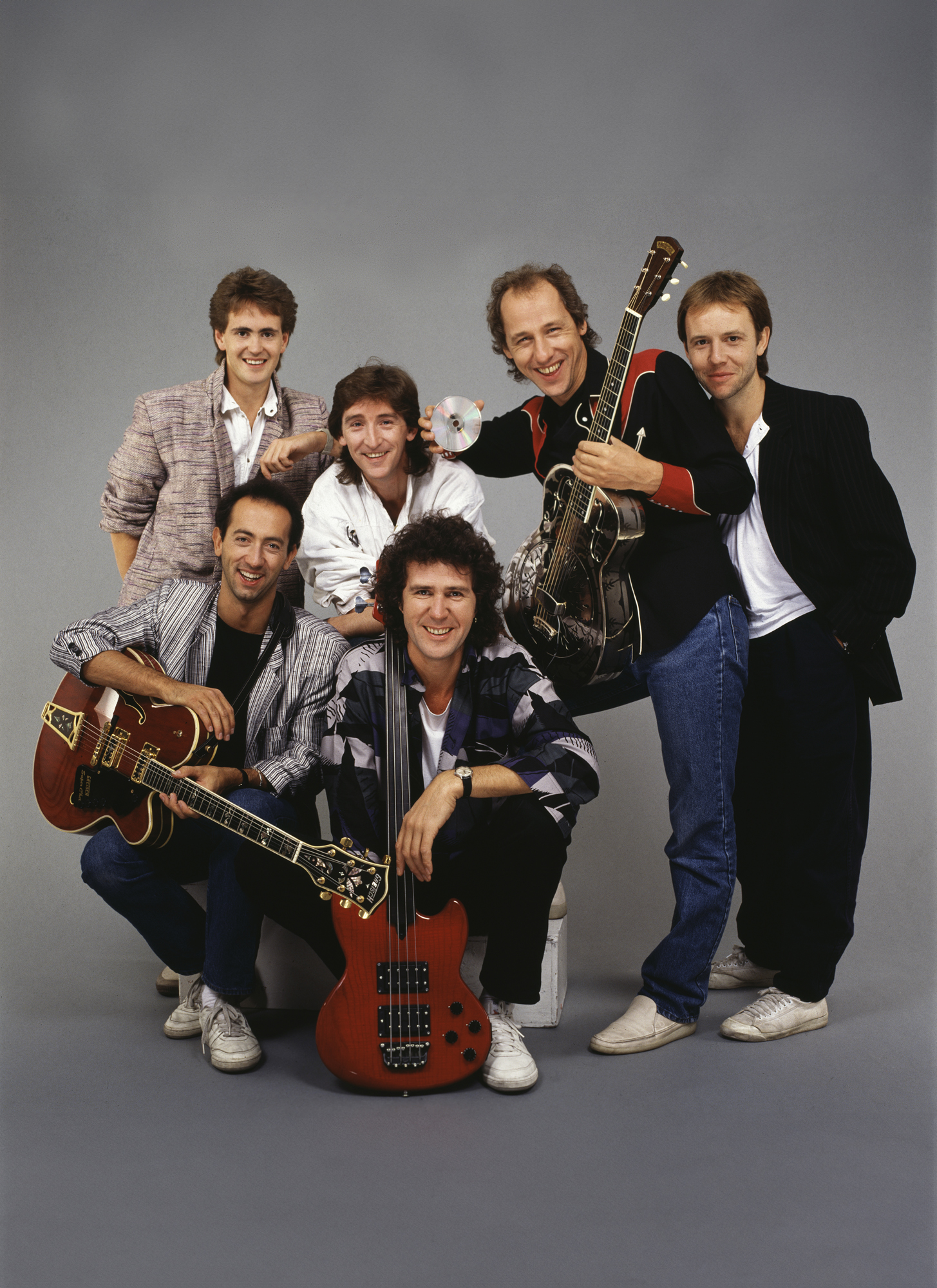 Dire Straits, New York, 1985. Pictured are, from left, Guy Fletcher (standing rear), Jack Sonni (sitting, with guitar), Terry Williams (in white jacket), John Illsley (sitting with bass guitar), Mark Knopfler (with acoustic guitar), and Alan Clark. Knopfler also holds a compact disc in one hand; his band's album, 'Brothers in Arms'