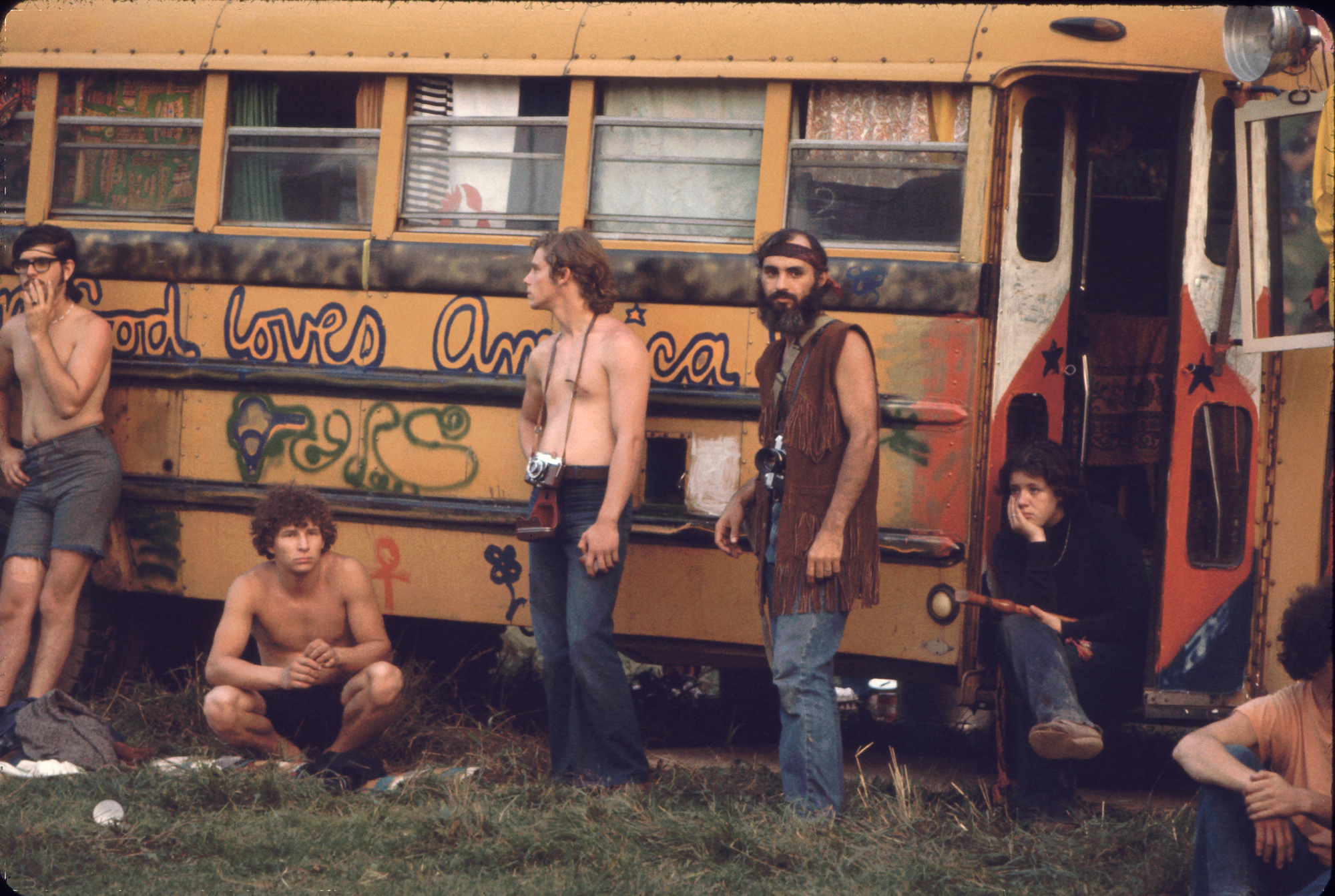 Hog Farm Bus At Woodstock Near the 'Free Stage' at the Woodstock Music and Arts Fair, several men, two with cameras around their necks, lean against a decorated school bus used by the Hog Farmers