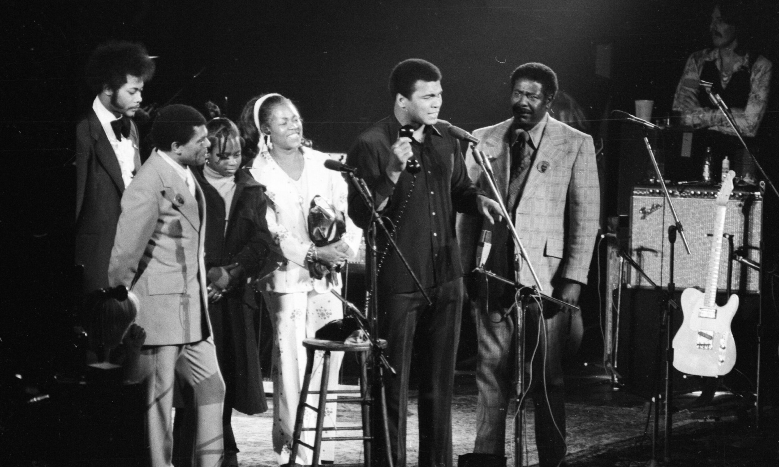 Muhammad Ali holds telephone during hookup with the State Prison at Clinton for conversation with Rubin (Hurricane) Carter. Mrs. Carter (in white), daughter and others listened during benefit Rolling Thunder Revue Concert given by Bob Dylan at Madison Square Garden.